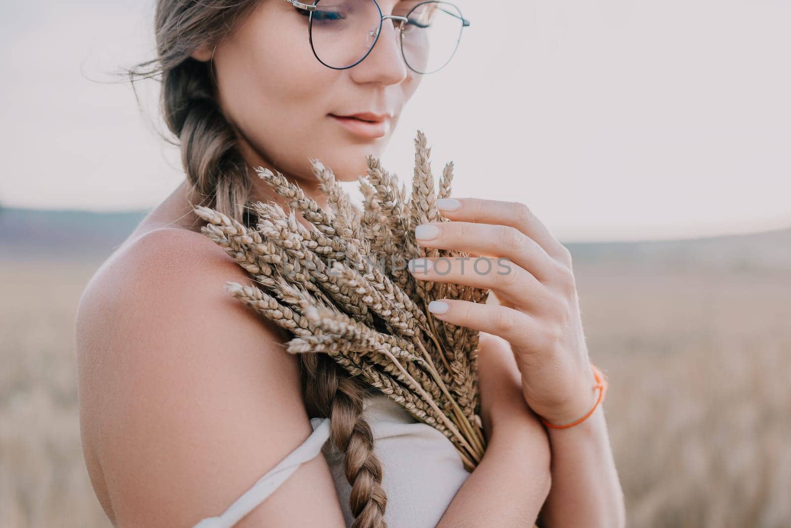A woman is in a wheat field holding a bunch of wheat close to her body, her arms crossed, wearing a white top and glasses, she is looking down at the wheat.