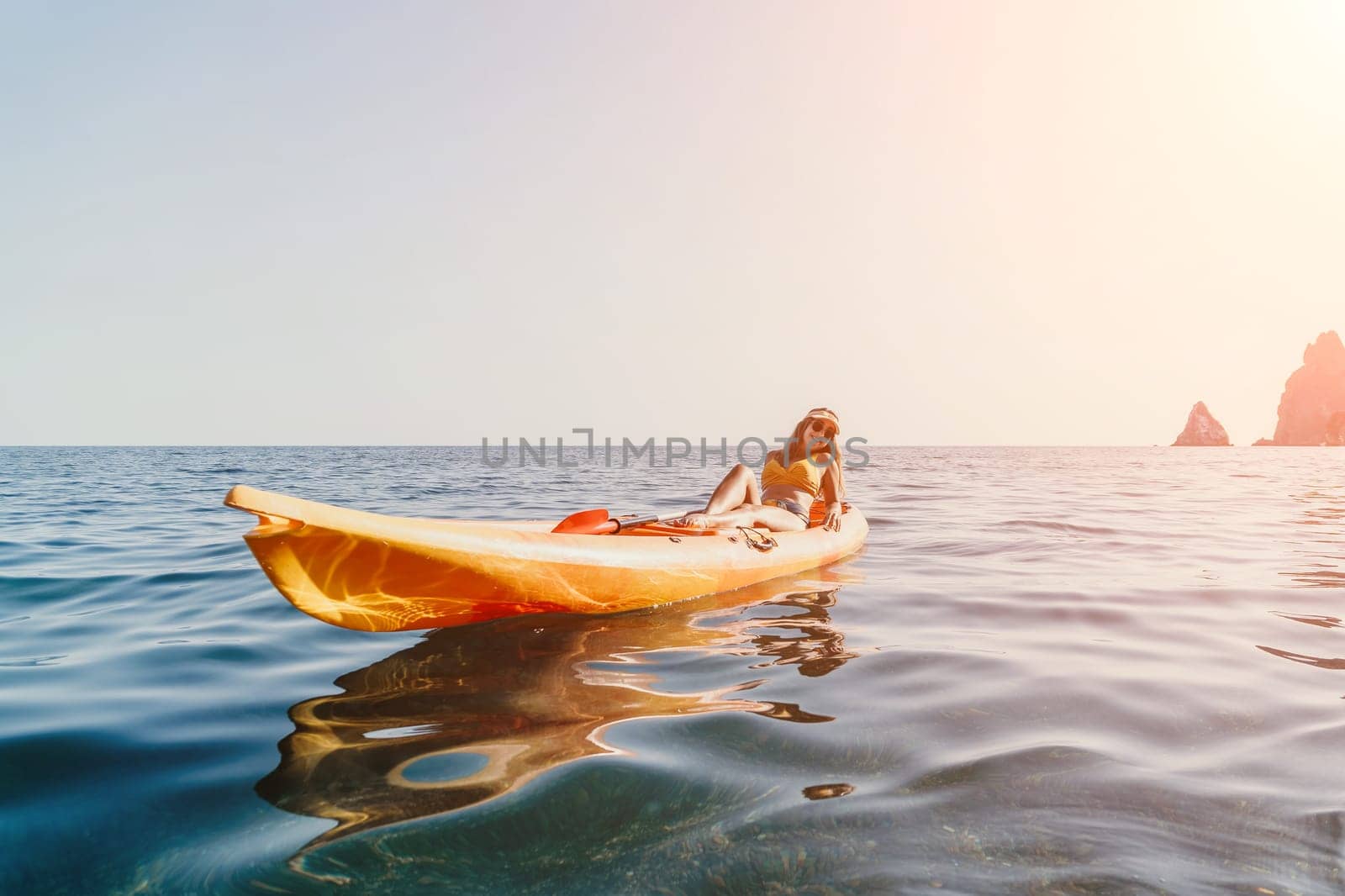 A woman kayaks on a calm sea with rocky cliffs in the distance. The sun is shining and the sky is blue.