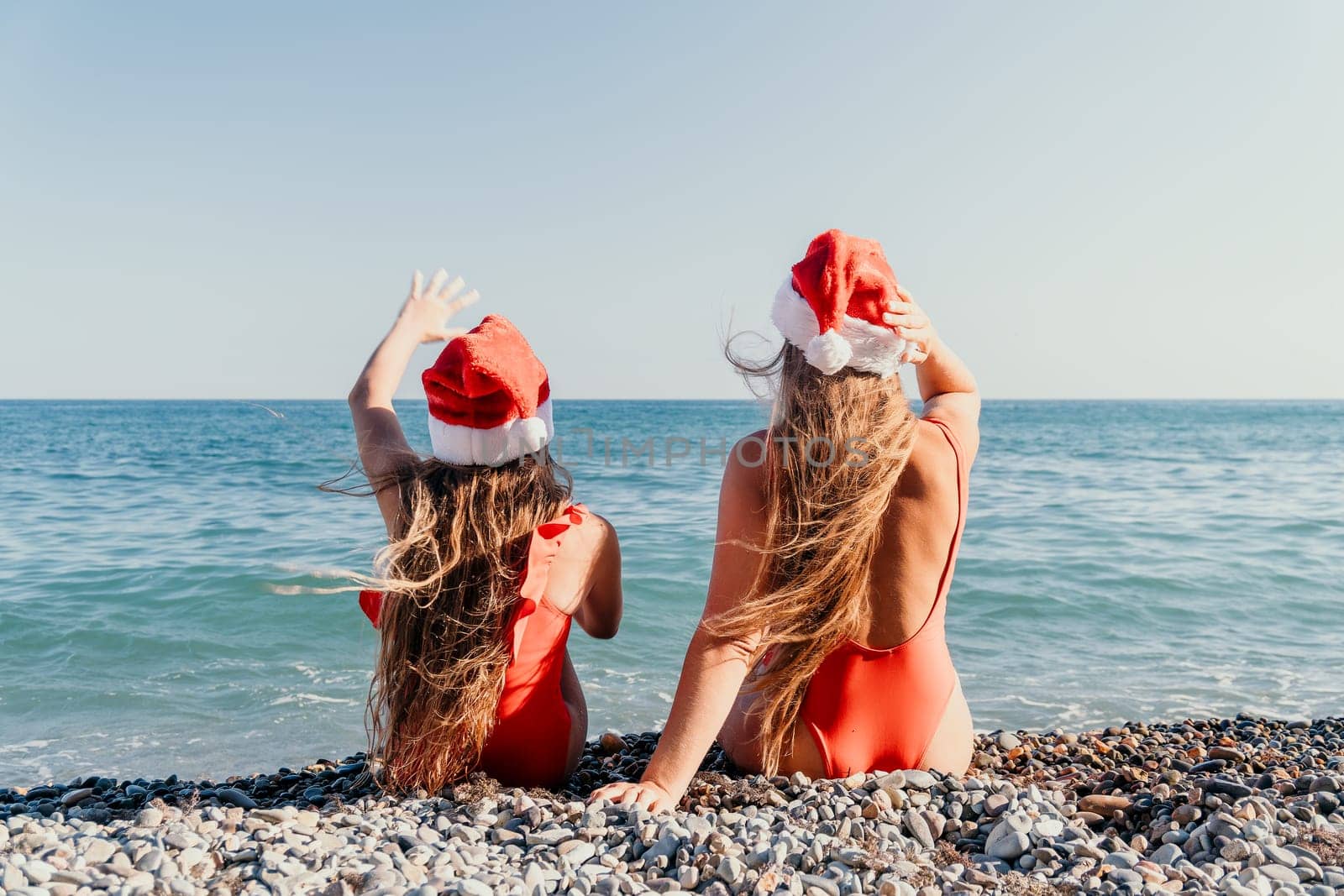 Two women in red swimsuits and Santa hats sit on a pebbled beach with the ocean in the background.