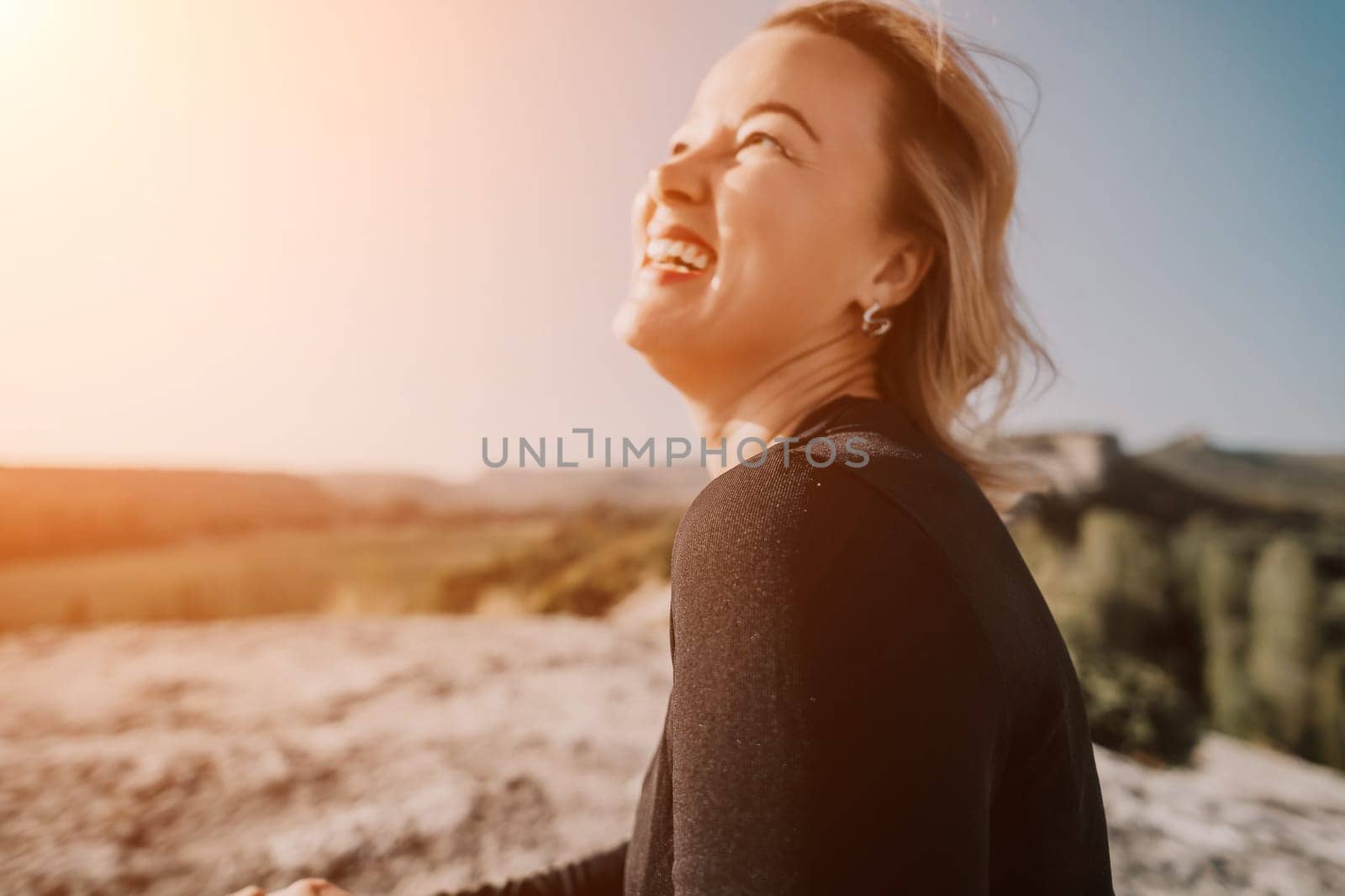 A woman with blonde hair laughs with her head thrown back while sitting on a rocky mountaintop, the landscape is blurred behind her.