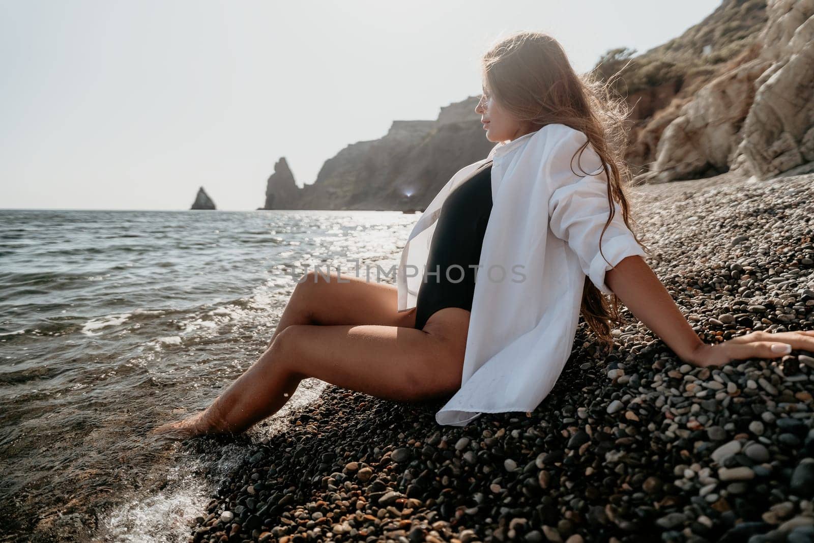 A woman wearing a white shirt and a black swimsuit sits on a pebbly beach, her feet in the water. The sea and distant rocky cliffs are in the background.