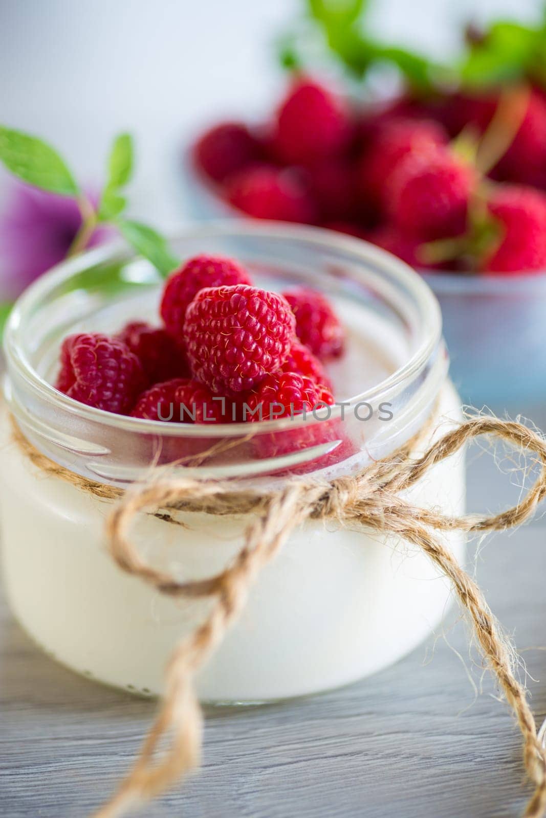 Sweet cooked homemade yogurt with fresh raspberries in a glass jar, on a light wooden table.