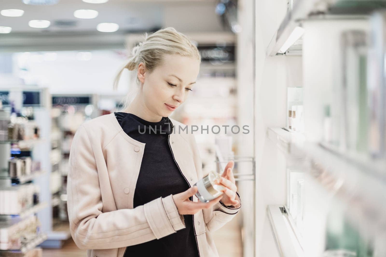 Elegant blond young woman choosing perfume in retail store. Beautiful blond lady testing and buying cosmetics in a beauty store.