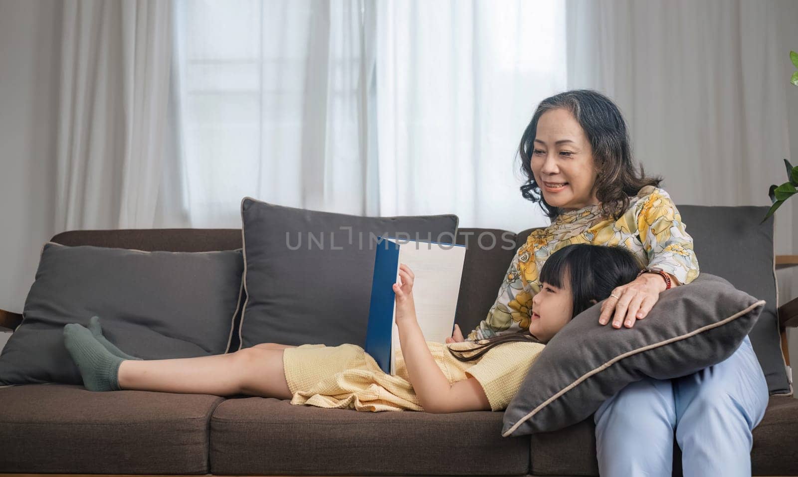 Grandma and granddaughter happily spend their free time reading books in the living room. An expression of love between an elderly woman and her adorable granddaughter..