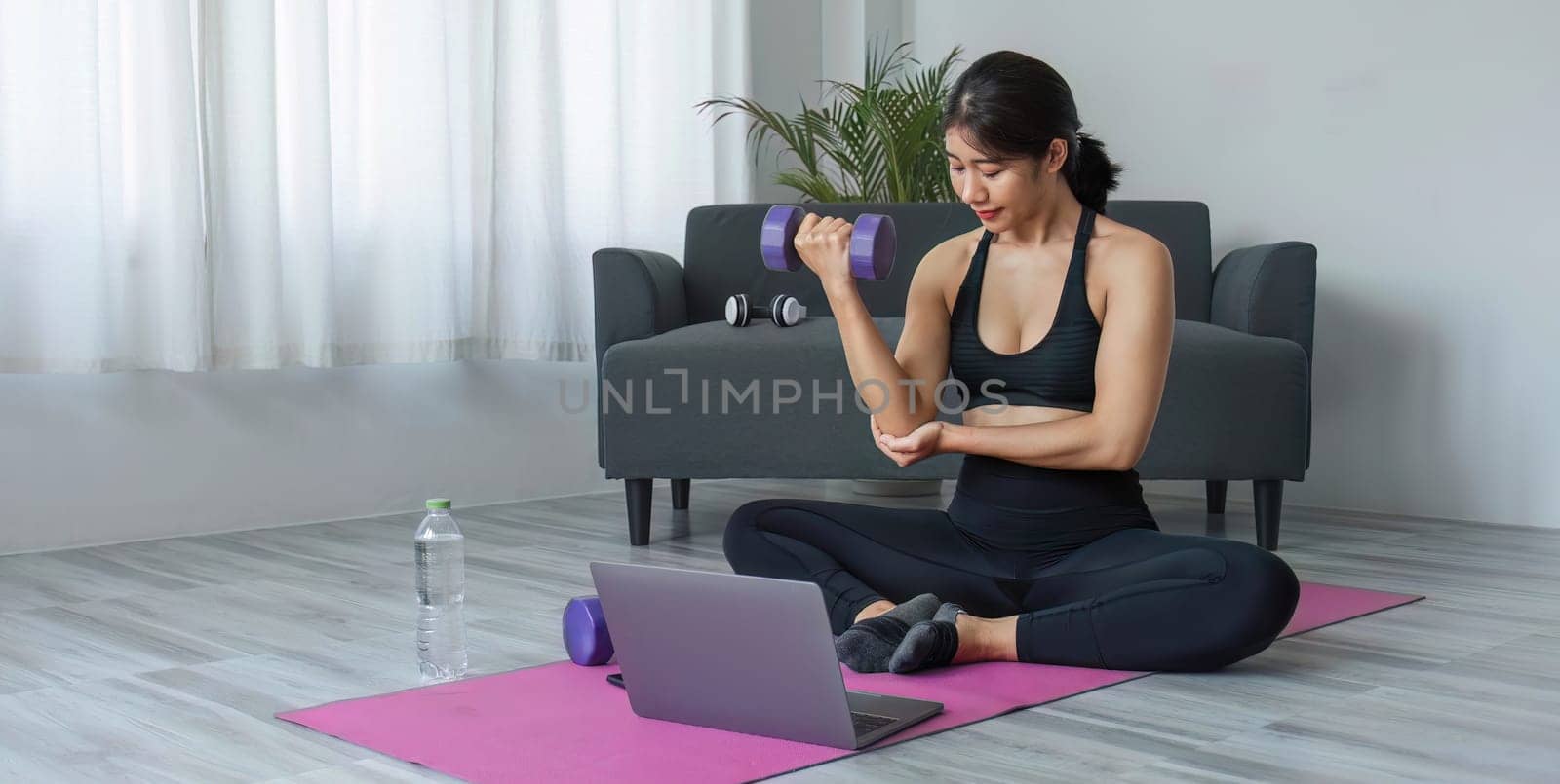 Young woman exercising in front of laptop Wear a sports bar outfit. Do yoga and lift dumbbells on the exercise mat. by wichayada