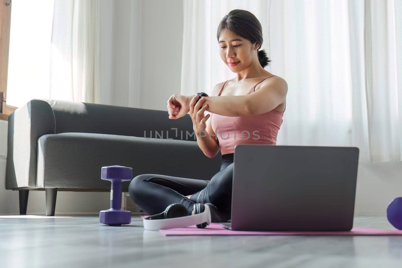 A young woman sits and looks at a smartwatch while exercising in front of her laptop. Wear a sports bar outfit. Do yoga and lift dumbbells on the exercise mat. by wichayada