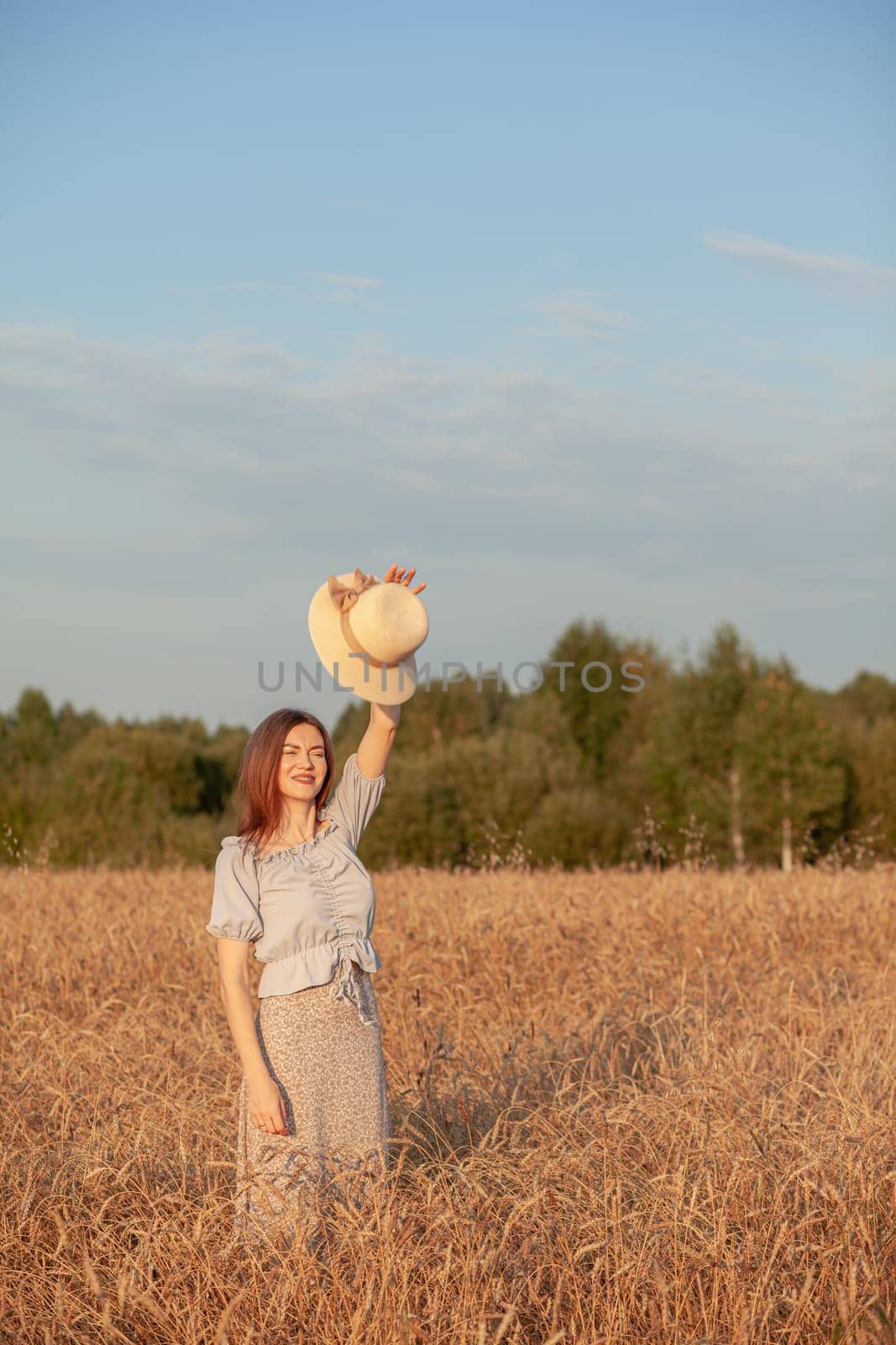 A young beautiful girl with braces on her teeth and long hair poses in a wheat field in the summer at sunset. The girl holds a hat in her hand against the background of a wheat field.