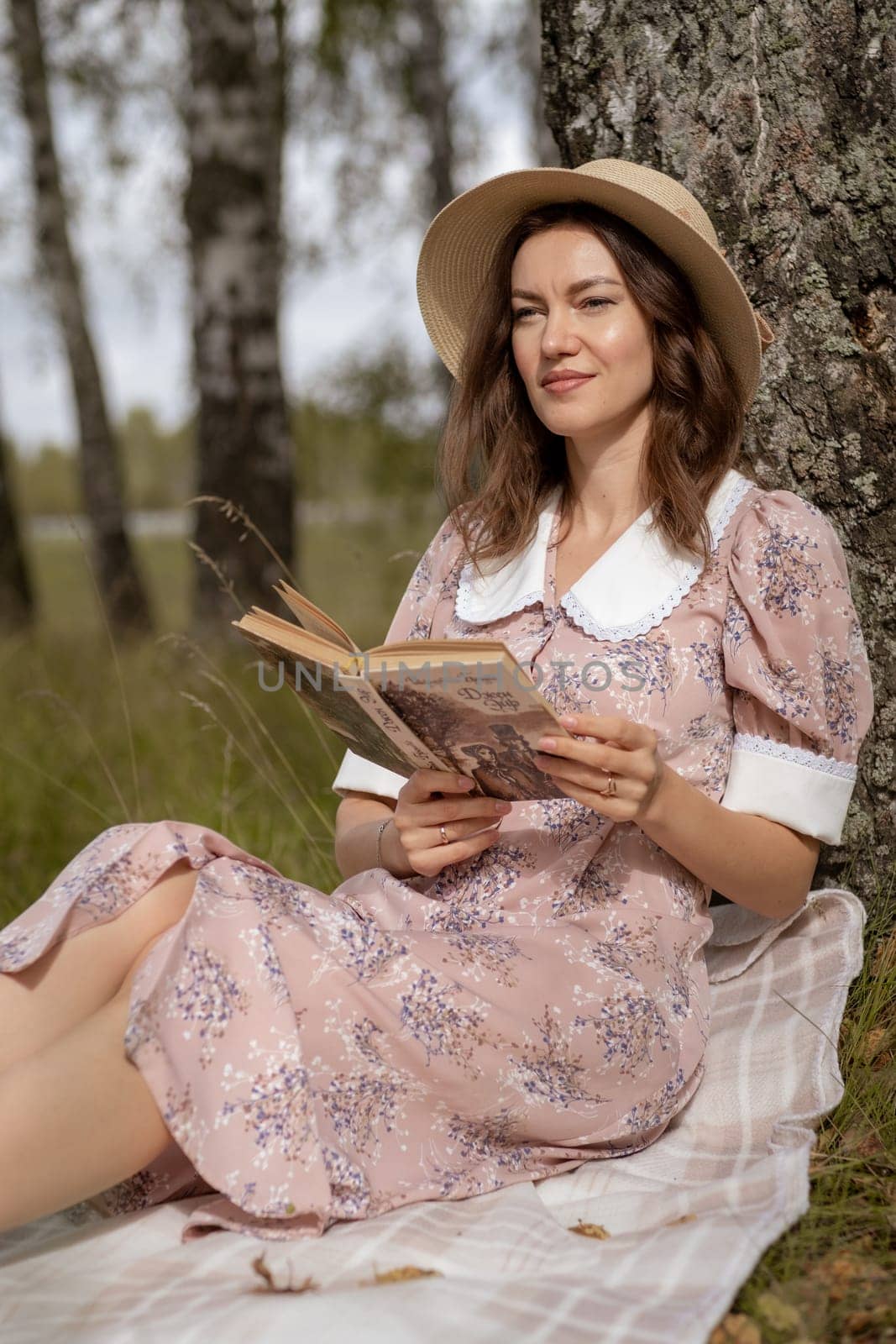 A young beautiful woman in a dress and a round hat reads a book outdoors in the forest and drinks tea. Romantic and vintage photo of a beautiful girl. Reading and relaxation