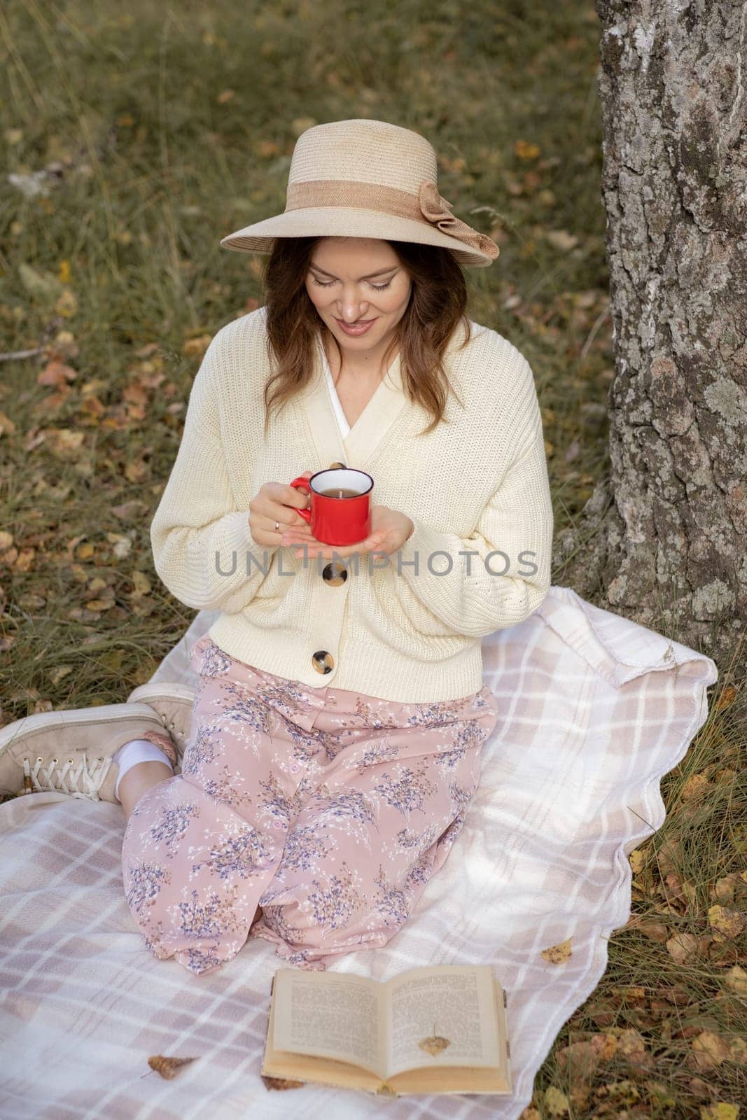 A young beautiful woman in a dress and a round hat reads a book outdoors in the forest and drinks tea. Romantic and vintage photo of a beautiful girl. Reading and relaxation