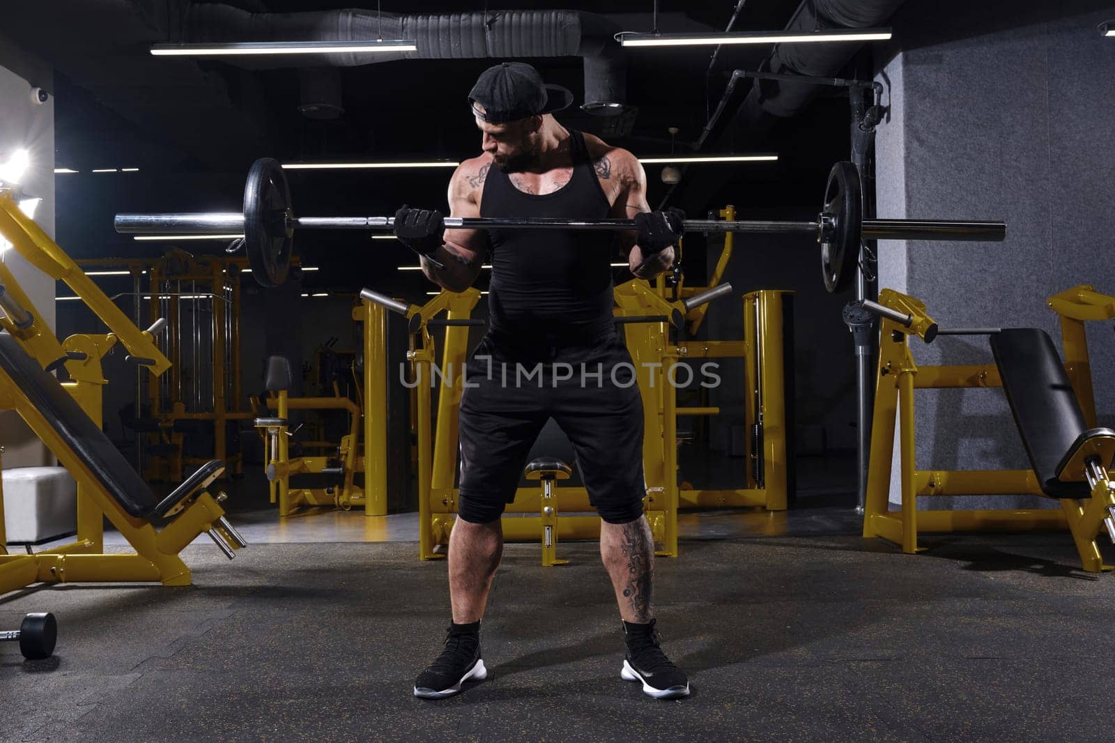 Attractive, tattooed, bearded sportsman in black sport gloves, shorts, vest, cap and sneakers. He is lifting a barbell, training his muscles, posing in dark gym with yellow equipment. Full length