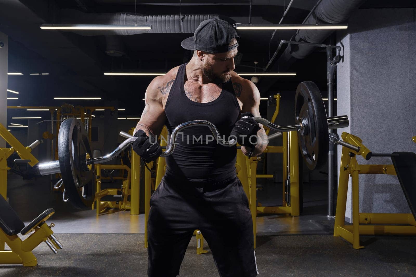 Tattooed, bearded sportsman in black sport gloves, shorts, vest and cap. He is lifting a barbell, training his biceps, looking at his muscles while posing in dark gym with yellow equipment. Close up