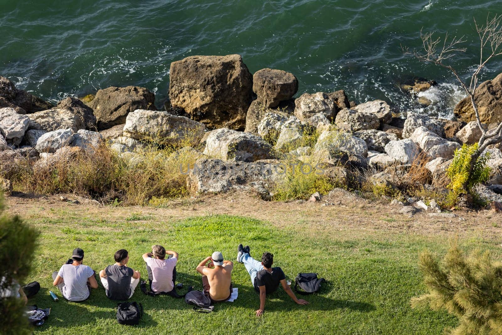 Young men sitting on the grass on the cliff by the sea. Mid shot