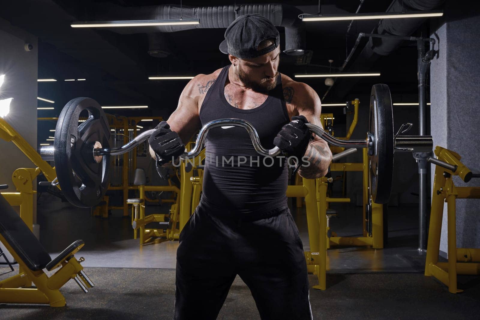 Tattooed, bearded man in black sport gloves, shorts, vest and cap. He is lifting a barbell, training his muscles, looking at his biceps while standing in dark gym with yellow equipment. Close up
