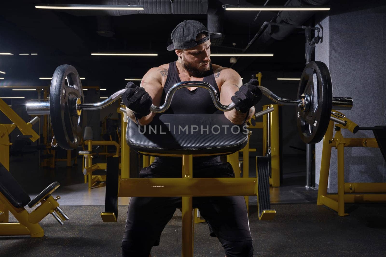 Tattooed, bearded athlete in black sport gloves, shorts, vest and cap. He is lifting a barbell, training his biceps while sitting on preacher curl bench at dark gym with yellow equipment. Close up
