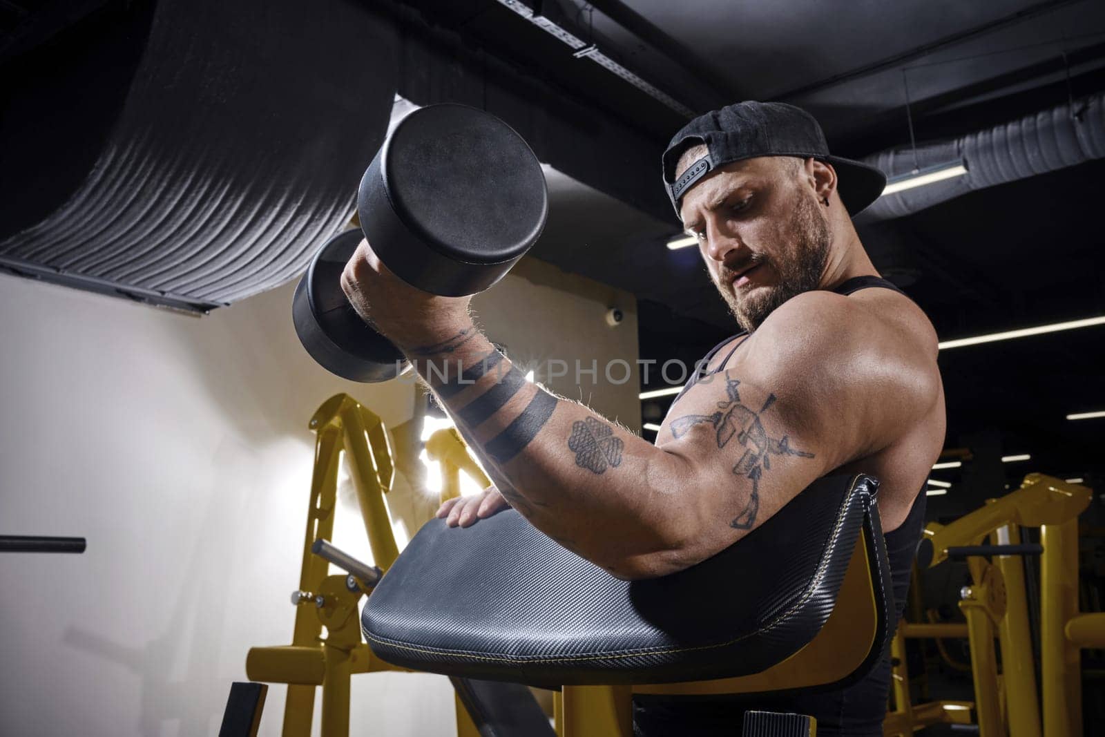 Tattooed guy with earring, in black vest and cap. He is lifting a dumbbell, training his biceps, sitting on preacher curl bench at dark gym. Close up by nazarovsergey