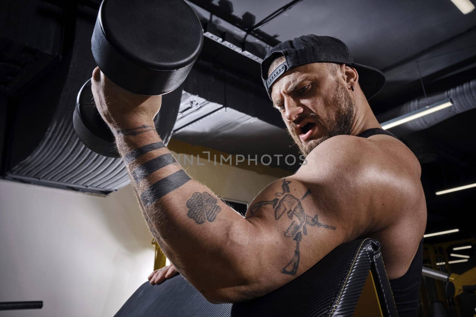 Tattooed, bearded man in black vest and cap. He is lifting a dumbbell, training his biceps, sitting on preacher curl bench at dark gym with yellow equipment. Sport, fitness. Close up, side view