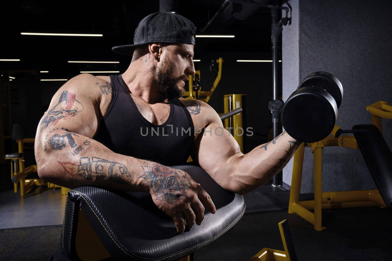 Tattooed, bearded guy with strong body, in black vest and cap. He is lifting a dumbbell, training his biceps, sitting on preacher curl bench at dark gym with yellow equipment. Sport, fitness. Close up