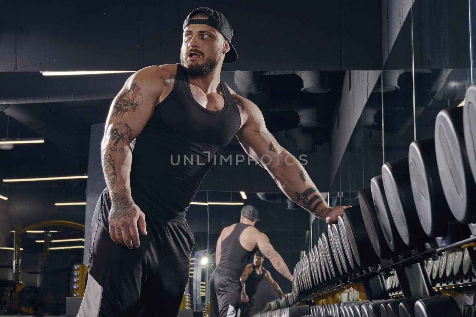 Young, tattooed, bearded male in black shorts, vest, cap. Going to do exercises with dumbbells for training muscles, leaning on set of black weights in dark gym. Sport, fitness. Close up, side view