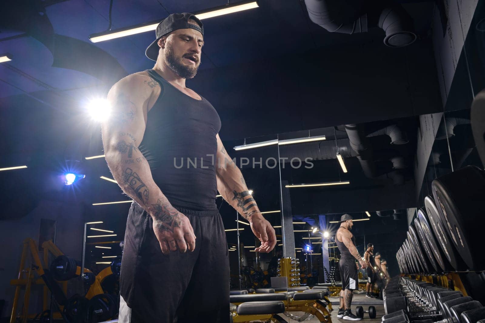 Tattooed, bearded athlete in black shorts, vest, cap. Going to do exercises with dumbbells for training his muscles, looking at set of black weights. Dark gym. Sport, fitness. Close up, side view