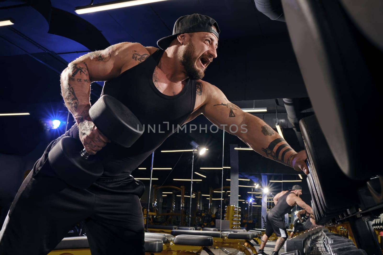 Handsome, tattooed, bearded muscular man in black shorts, vest, cap. Exercising with dumbbells for training his triceps, leaning on set of black weights. Dark gym. Sport, fitness. Close up, side view