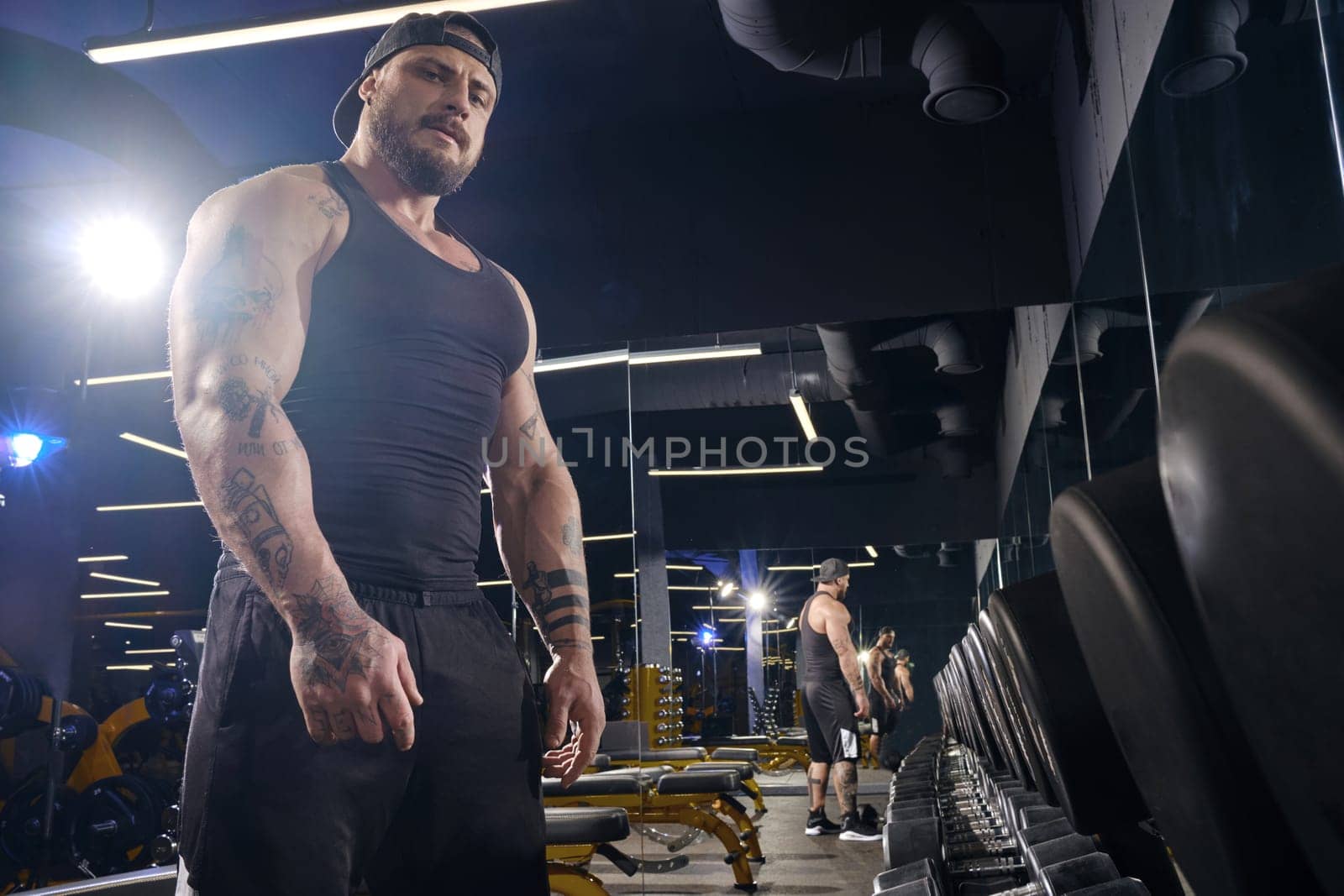 Tattooed, muscular bearded guy in black shorts, vest, cap. Looking at you, standing near set of black dumbbells. Dark gym. Sport. Close up by nazarovsergey