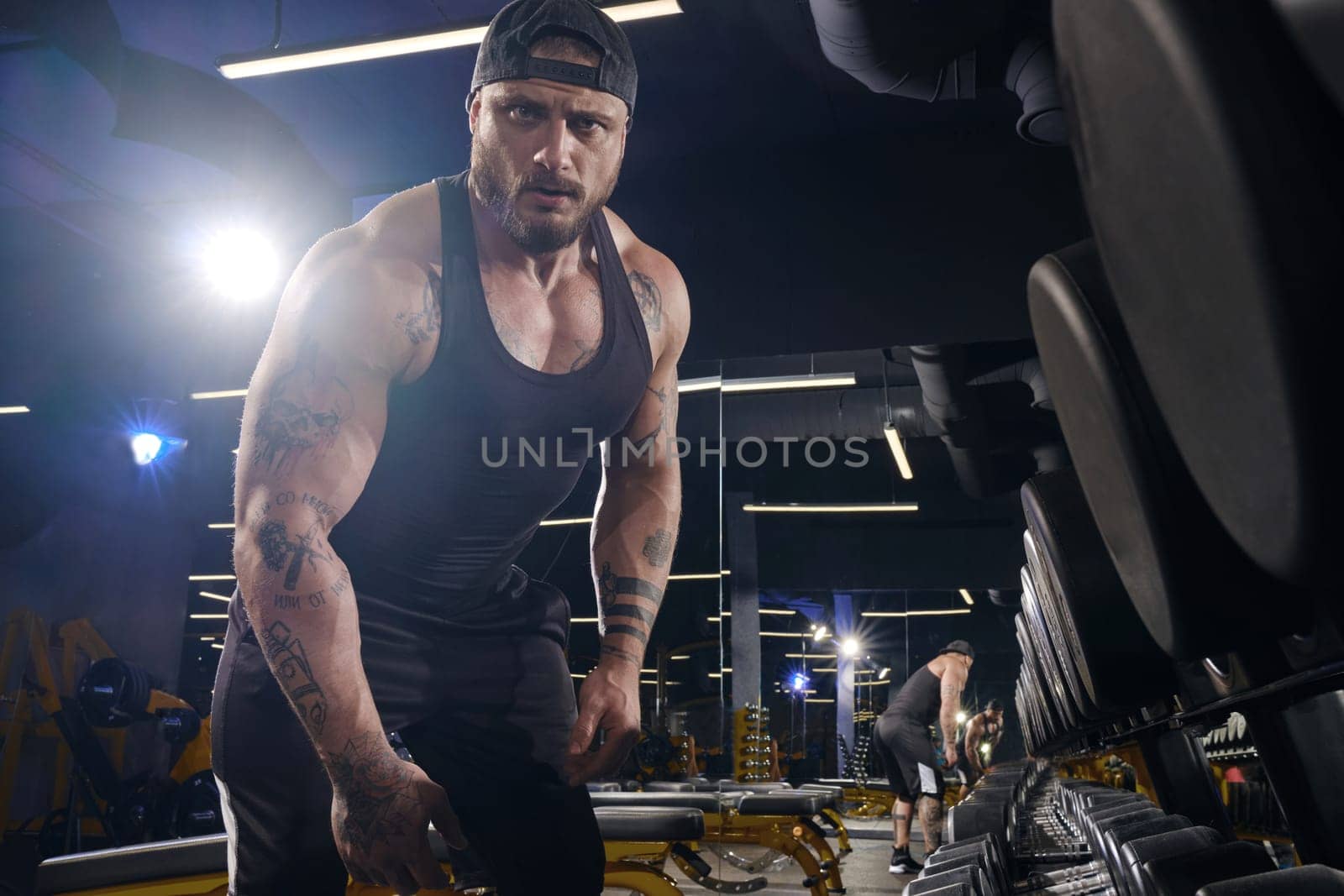 Handsome, tattooed, bearded sportsman in black shorts, vest, cap. Looking at you while standing near set of black dumbbells. Dark gym. Close up by nazarovsergey