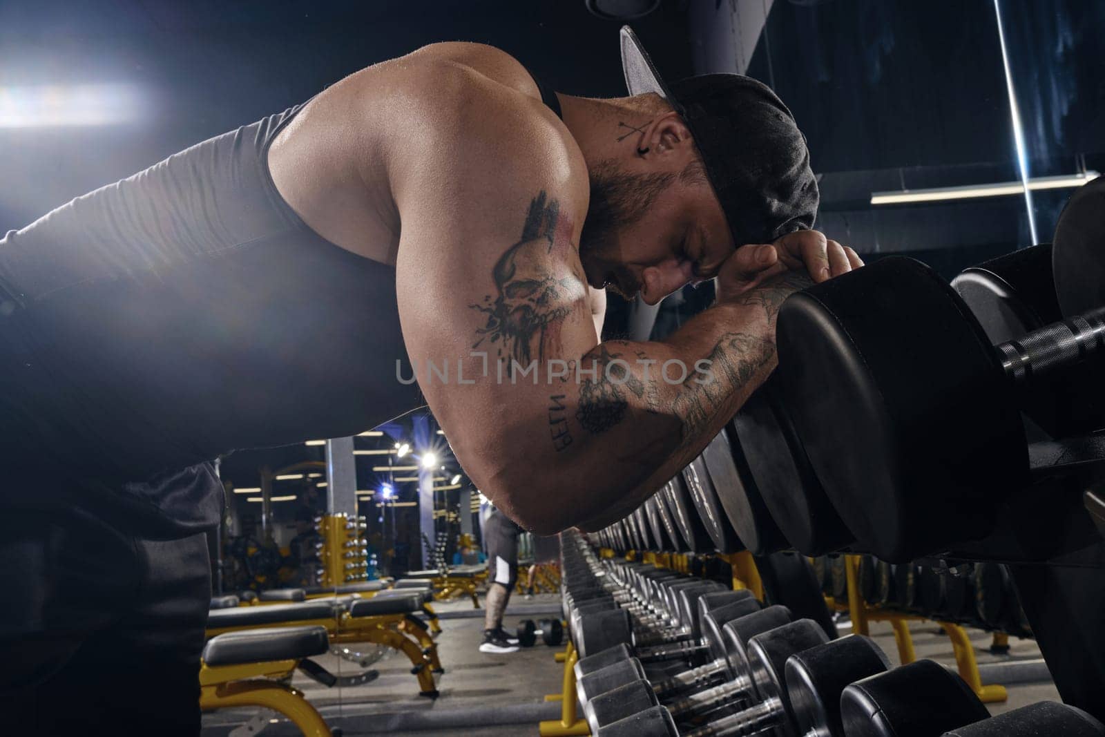 Handsome, tattooed, bearded muscular athlete in black shorts, vest, cap. Having rest leaning on set of black dumbbells by his head and hands. Dark gym. Sport, fitness, lifestyle. Close up, side view