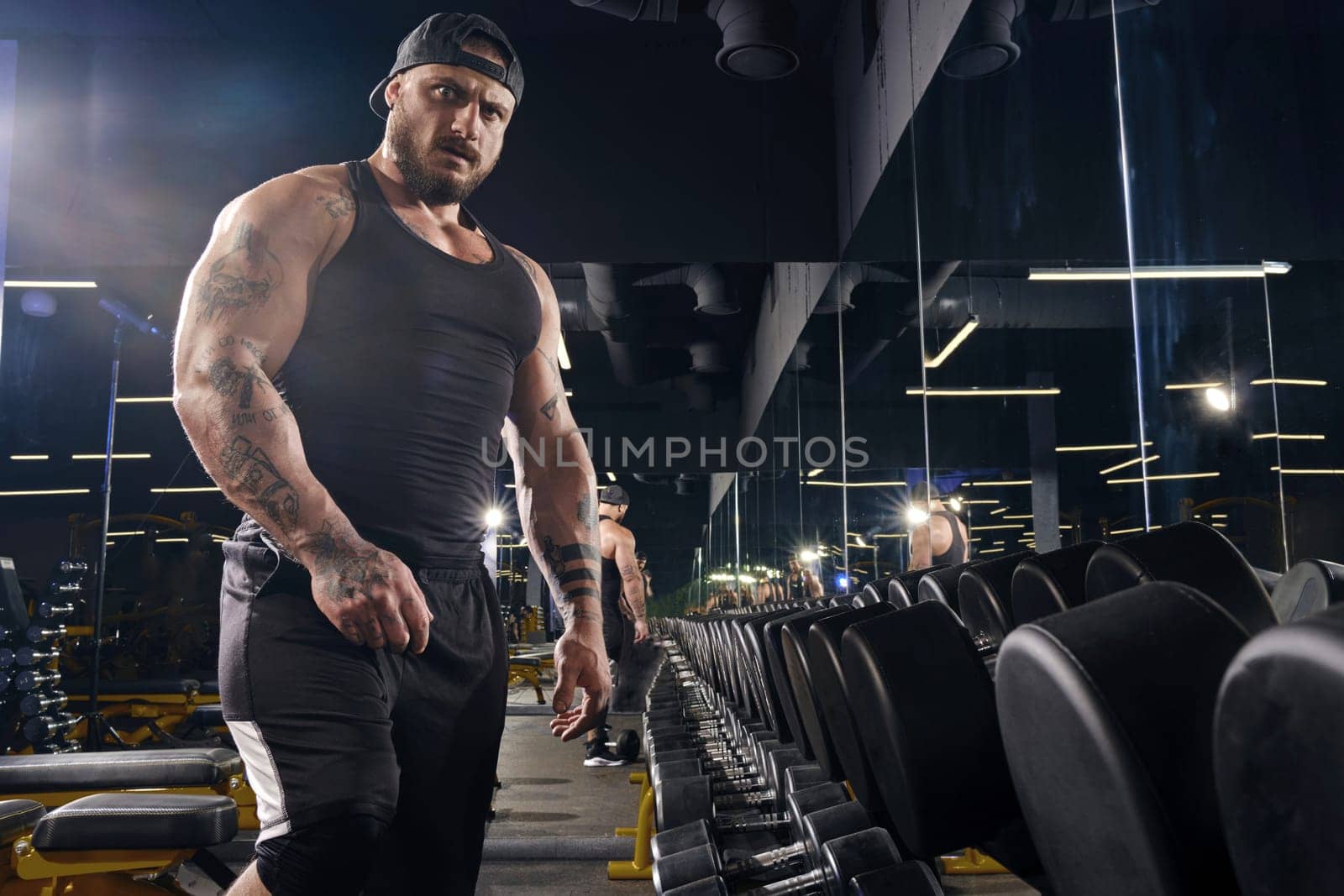 Attractive, tattooed, bearded man in black shorts, vest, cap. Going to do exercises with dumbbells for training his muscles, looking at you, standing near set of black weights. Dark gym. Close up