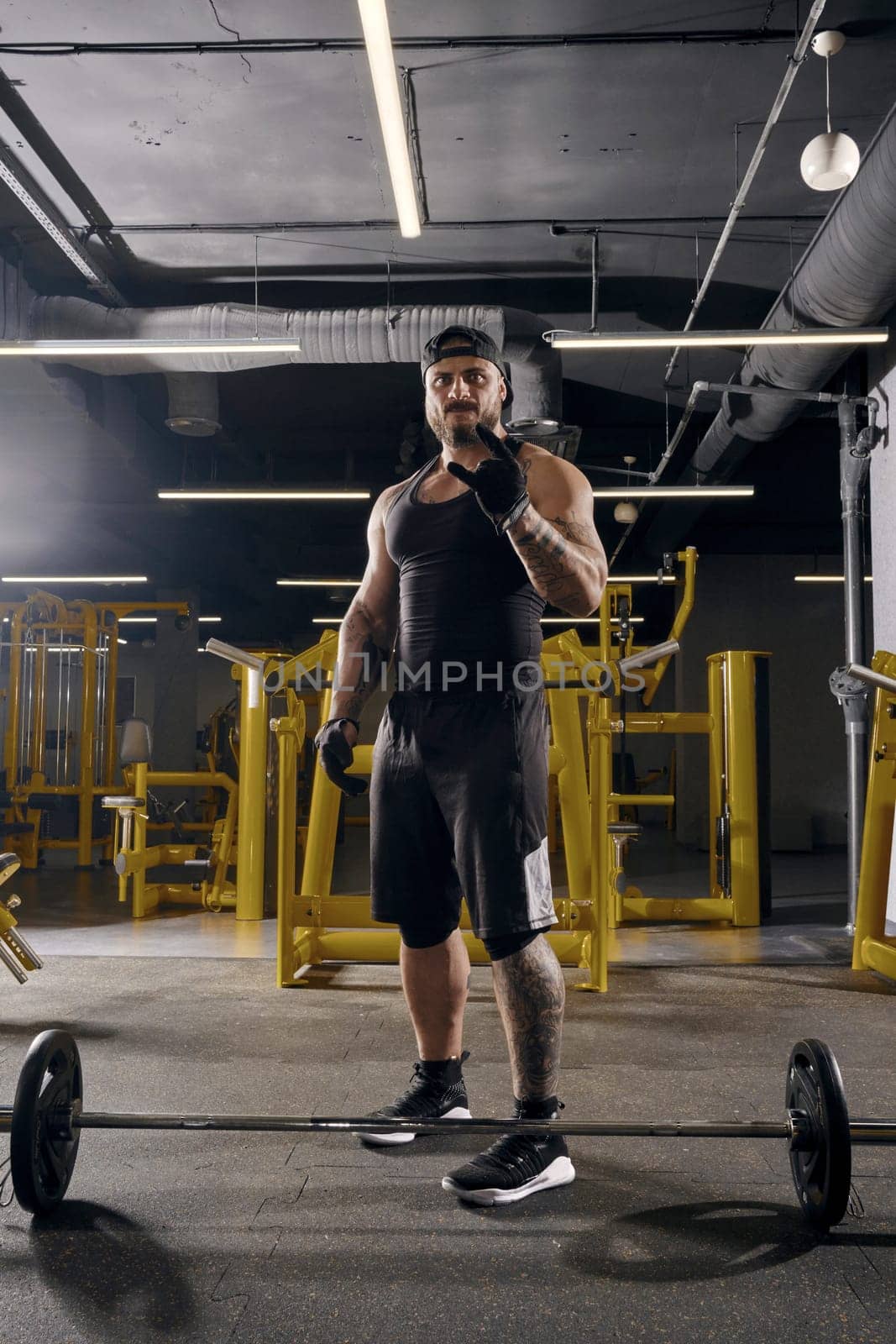 Attractive, tattooed, bearded man in black sport gloves, shorts, vest, cap and sneakers. Showing cool sign, barbell is on floor, going to do exercises for training his muscles. Dark gym. Full length