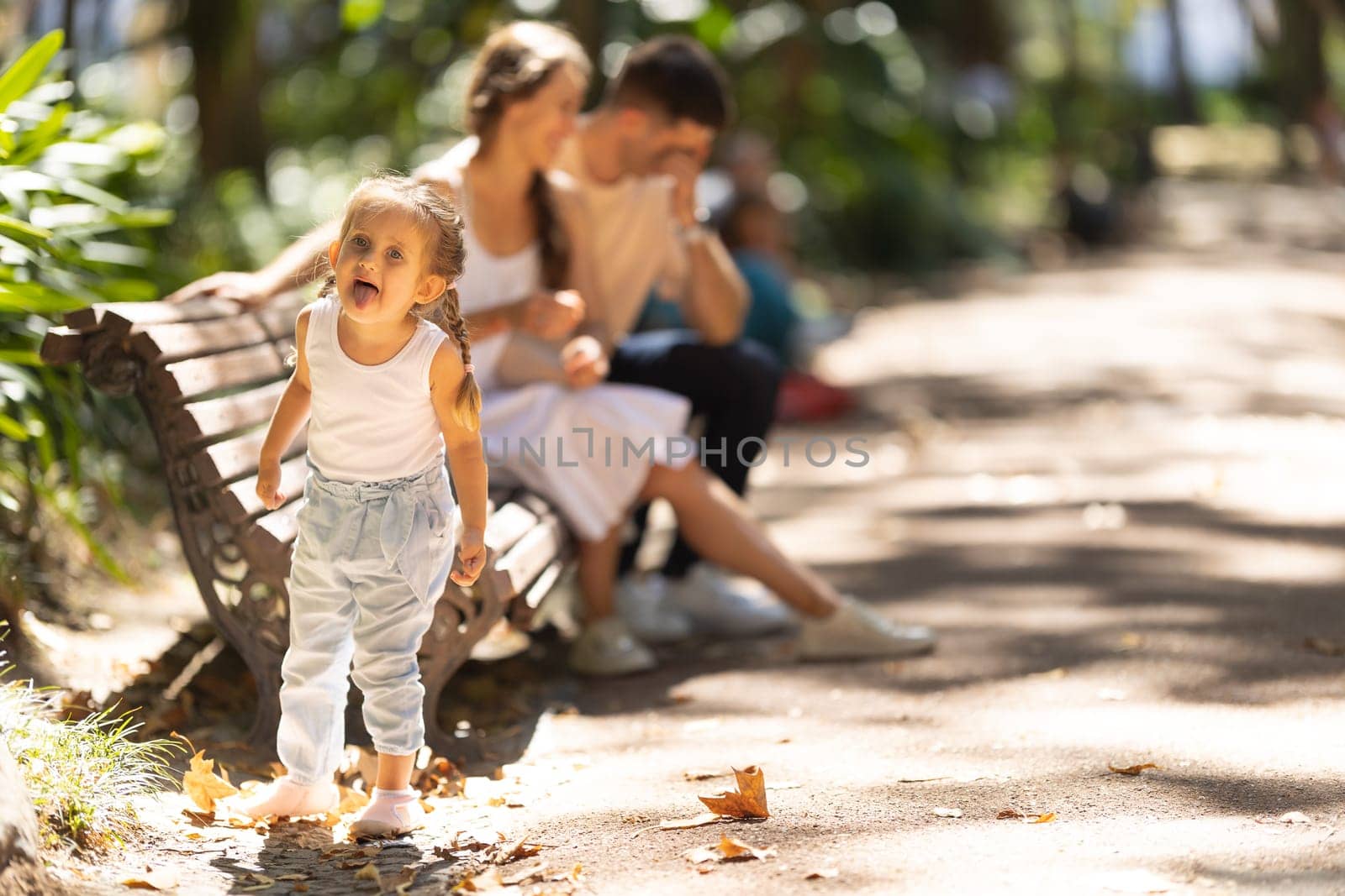 Family in the park - parents sitting on the bench and their little daughter showing tongue on the foreground. Mid shot