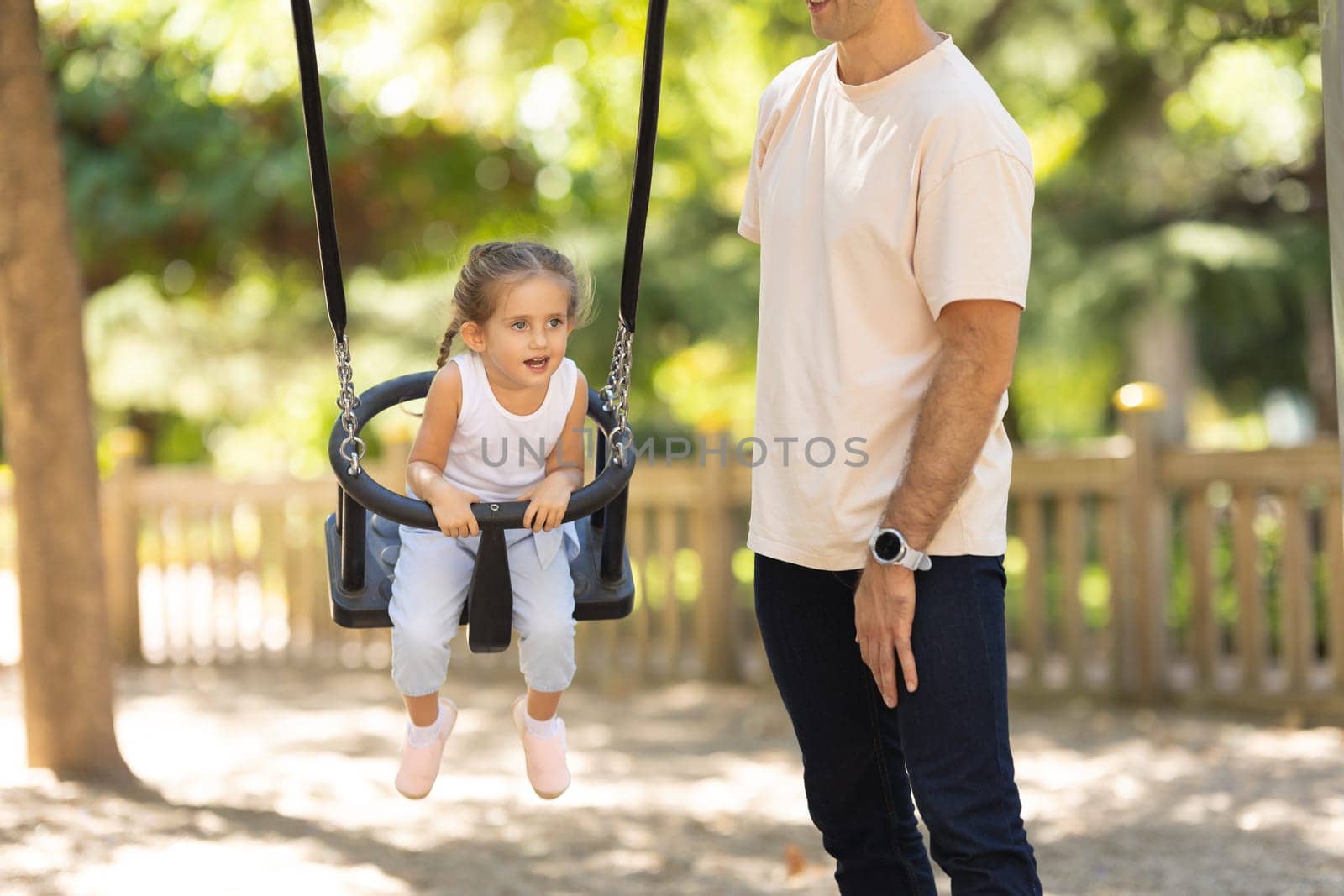 A man riding his little daughter on the swings in the summer park. Mid shot