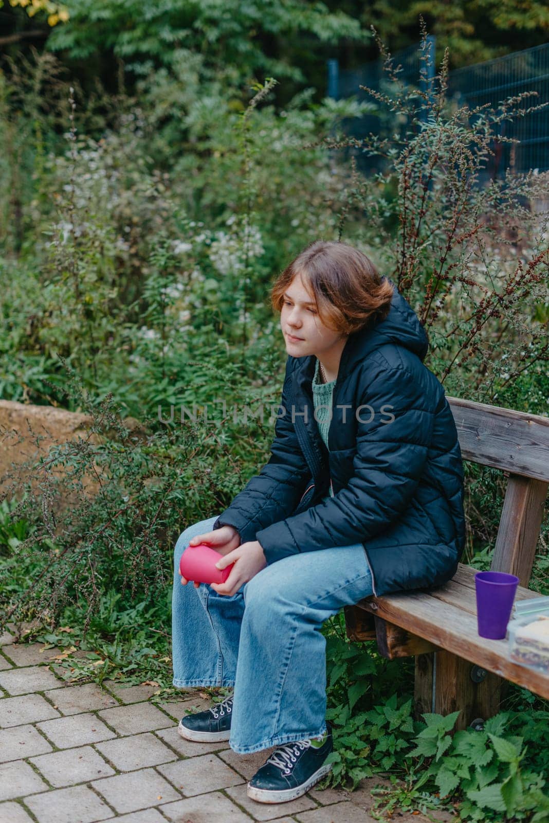 Blonde girl holding cup sitting on bench in the park. Autumn season. Young Teen Girl relaxing on bench with cup of coffee or tea.