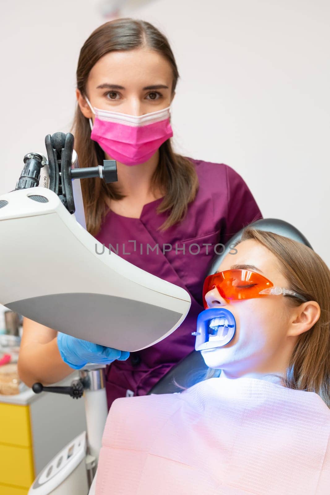 Young dentist performs bleaching of teeth using UV lamp for woman at dentist clinic, vertical photo