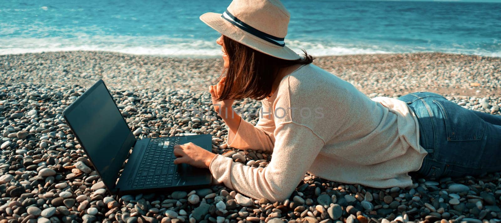 A young girl in a light hat and casual sweater lies on the beach by the sea with a laptop on a sunny day, works, studies, buys tickets during trip, a woman rests on vacation and types on the keyboard.