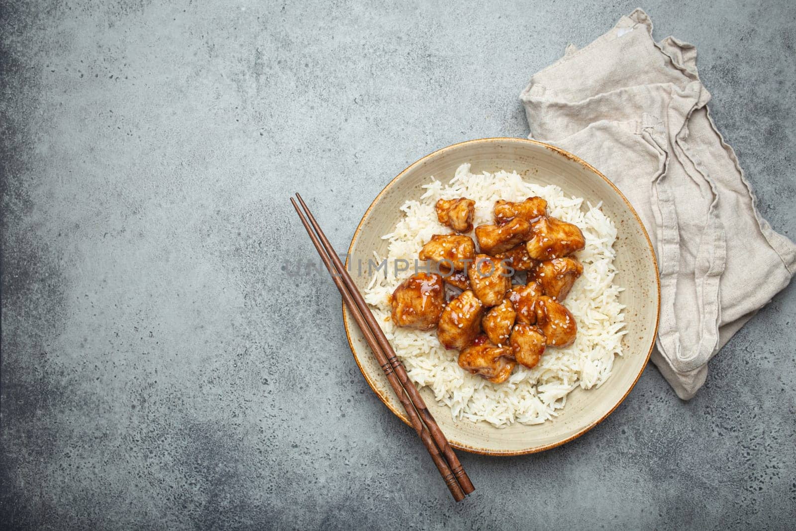 Chinese sweet and sour sticky chicken with sesame seeds and rice on ceramic plate with chopsticks top view on gray rustic stone background, traditional dish of China. Copy space by its_al_dente