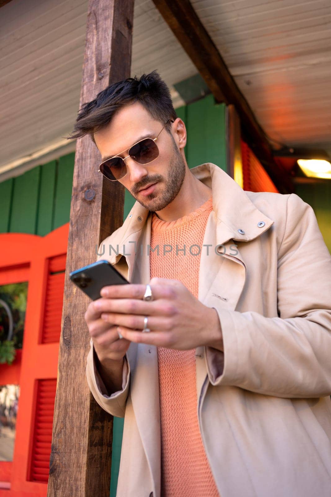 Male adult wearing sunglasses using smartphone for sending message. Man in stylish trench coat and fashionable eyewear texting on cellphone. Modern lifestyle, connection concept