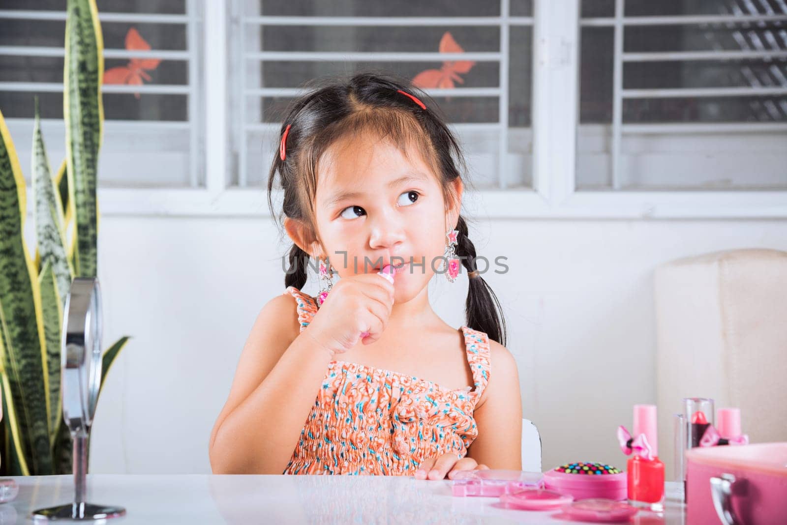 little girl making makeup her face she look in the mirror and applying red lipstick to mouth by Sorapop