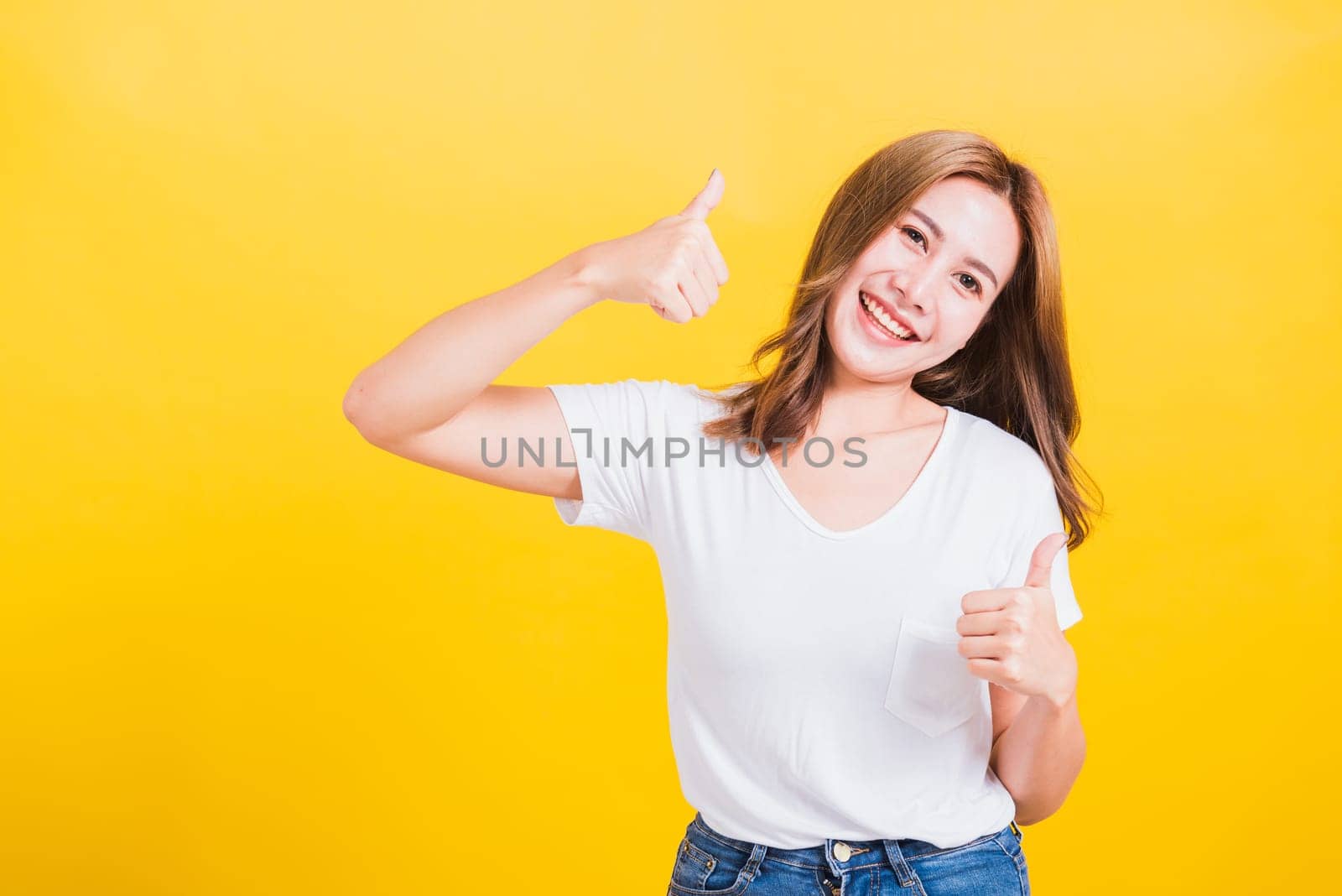 Portrait Asian Thai beautiful happy young woman smiling wear white t-shirt standing successful woman giving two thumbs up gesture sign in studio shot, isolated on yellow background with copy space