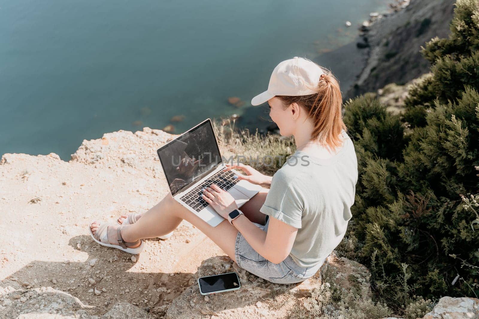 A young woman in a white cap sits on a cliff overlooking the ocean, working on her laptop.
