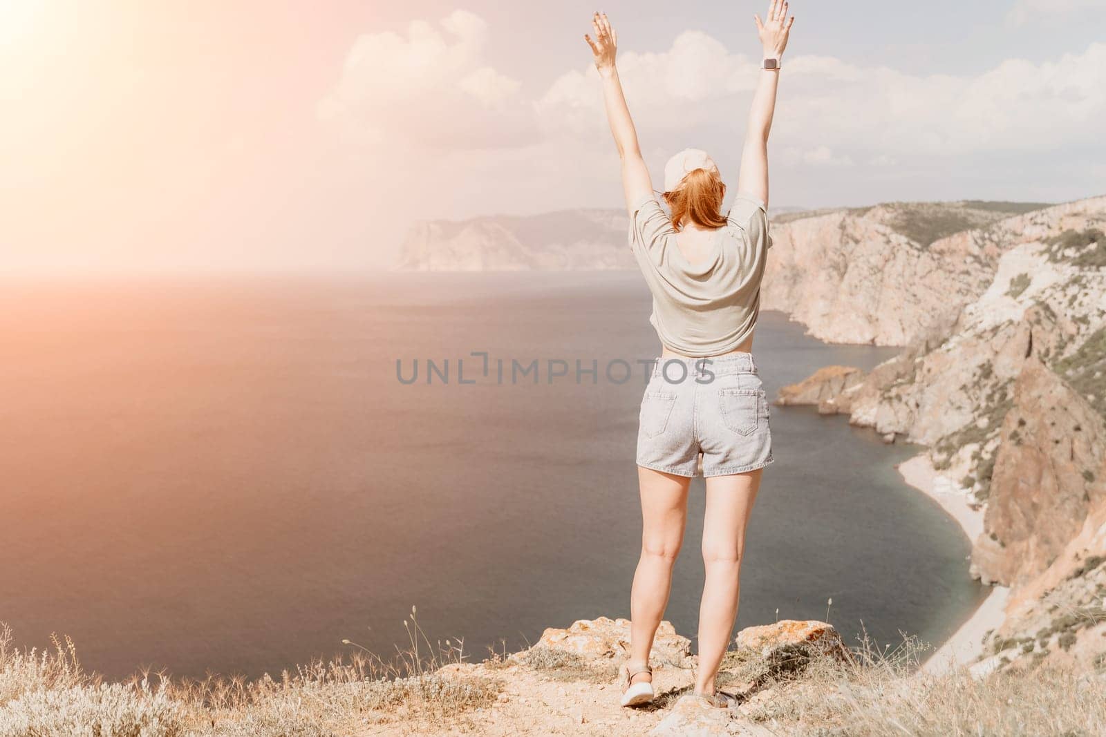 A woman in a casual outfit stands on a cliff, arms raised in celebration, with a beautiful seascape behind her.