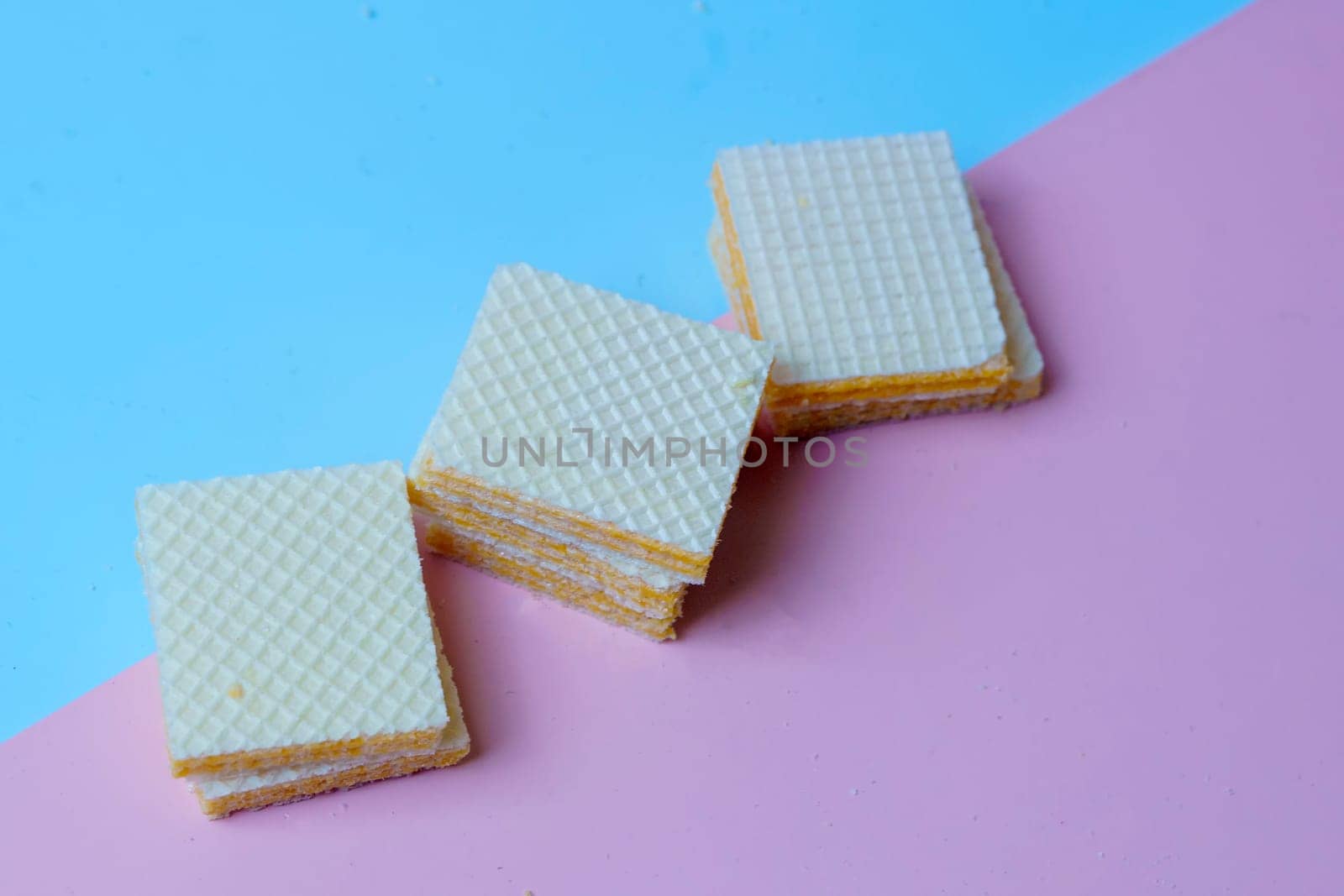 A pile of square, yellow wafers arranged on a blue and pink background. Copy space