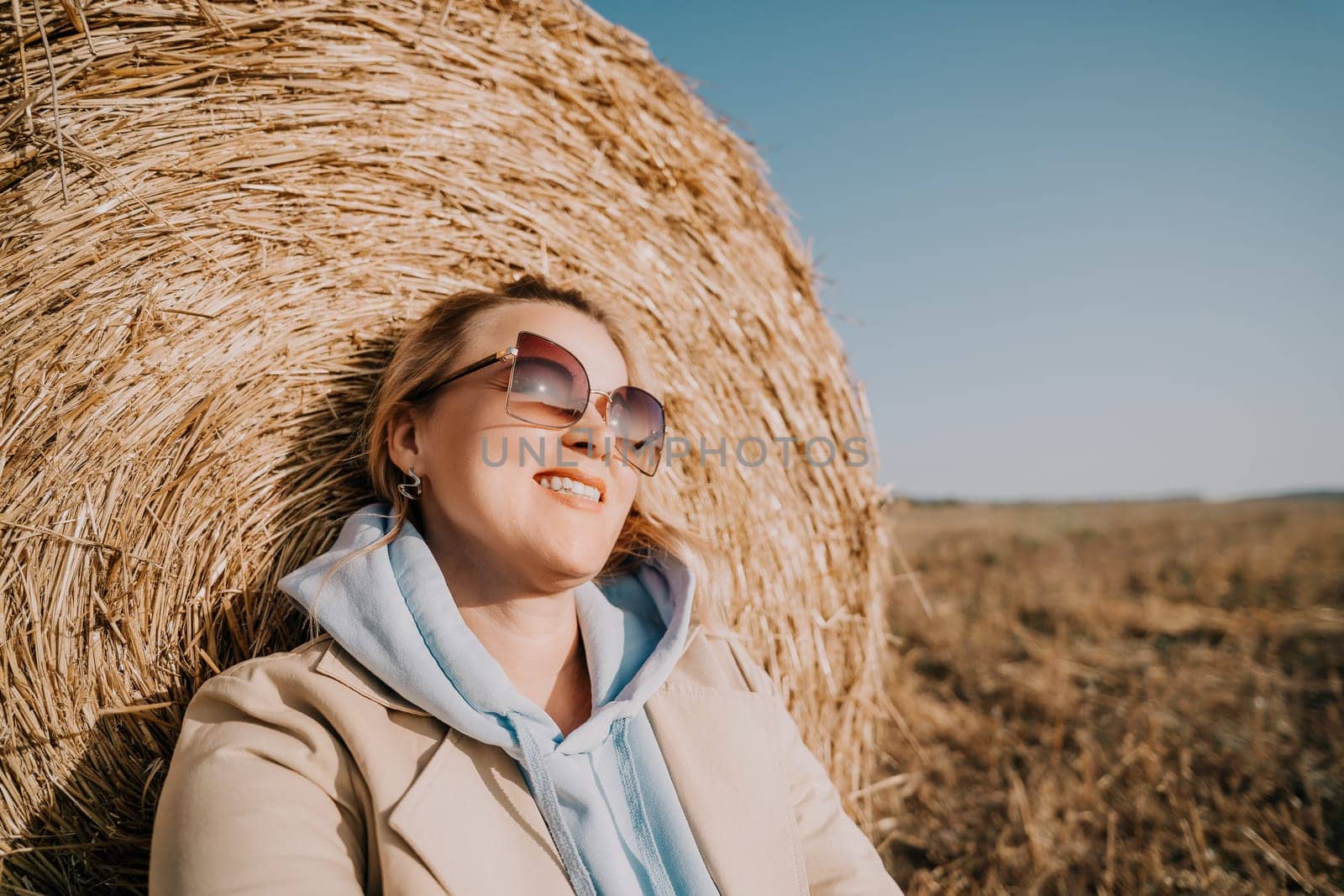 A woman wearing sunglasses and a beige jacket leans against a hay bale, smiling warmly.