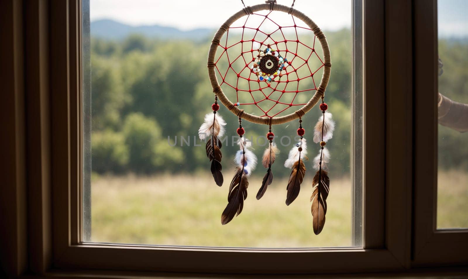 A dreamcatcher with red string and feathers hangs from a window. There is a view of green trees and grass beyond the window.
