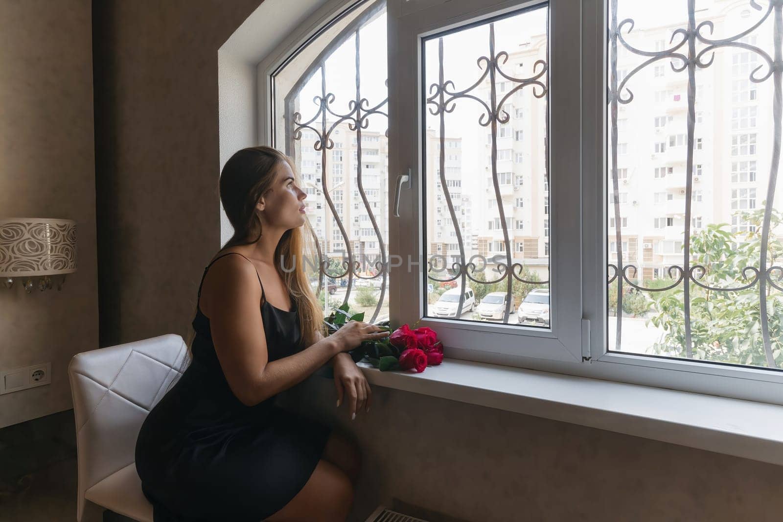A woman in a black dress is sitting in front of a window with a bouquet of red roses