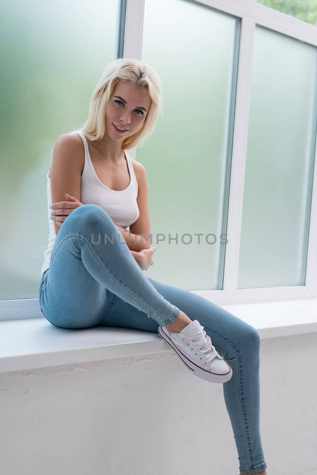 Young sexy blonde dressed in white top and jeans posing on the window sill in studio