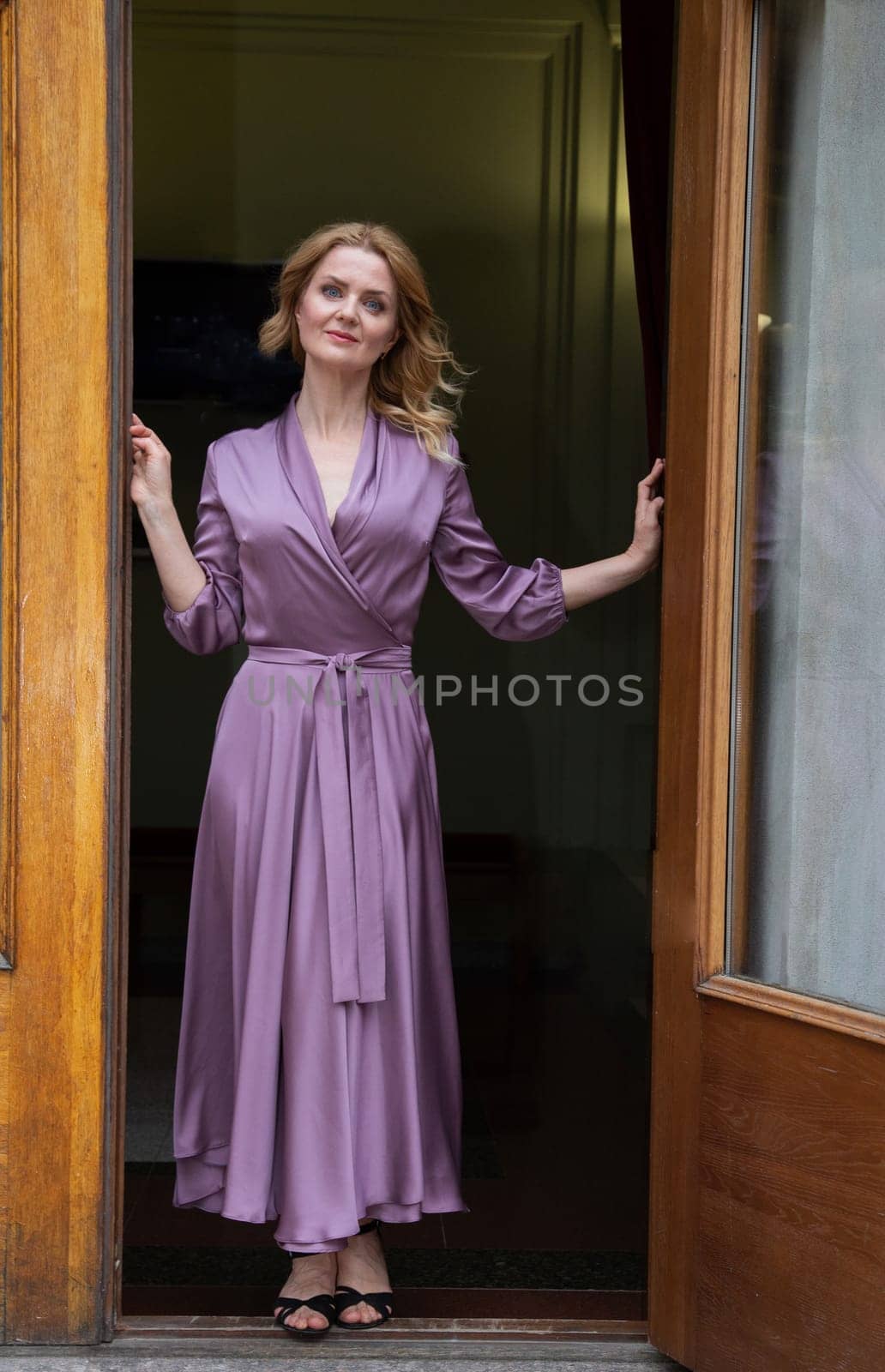 A pretty woman in a purple dress with beautiful yellow hair stands in a doorway against the backdrop of a dark room.