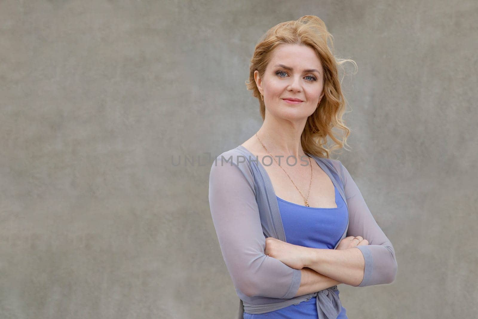 A beautiful woman in a good mood in a blue dress stands near the wall crossed, folded arms and looks at the camera.