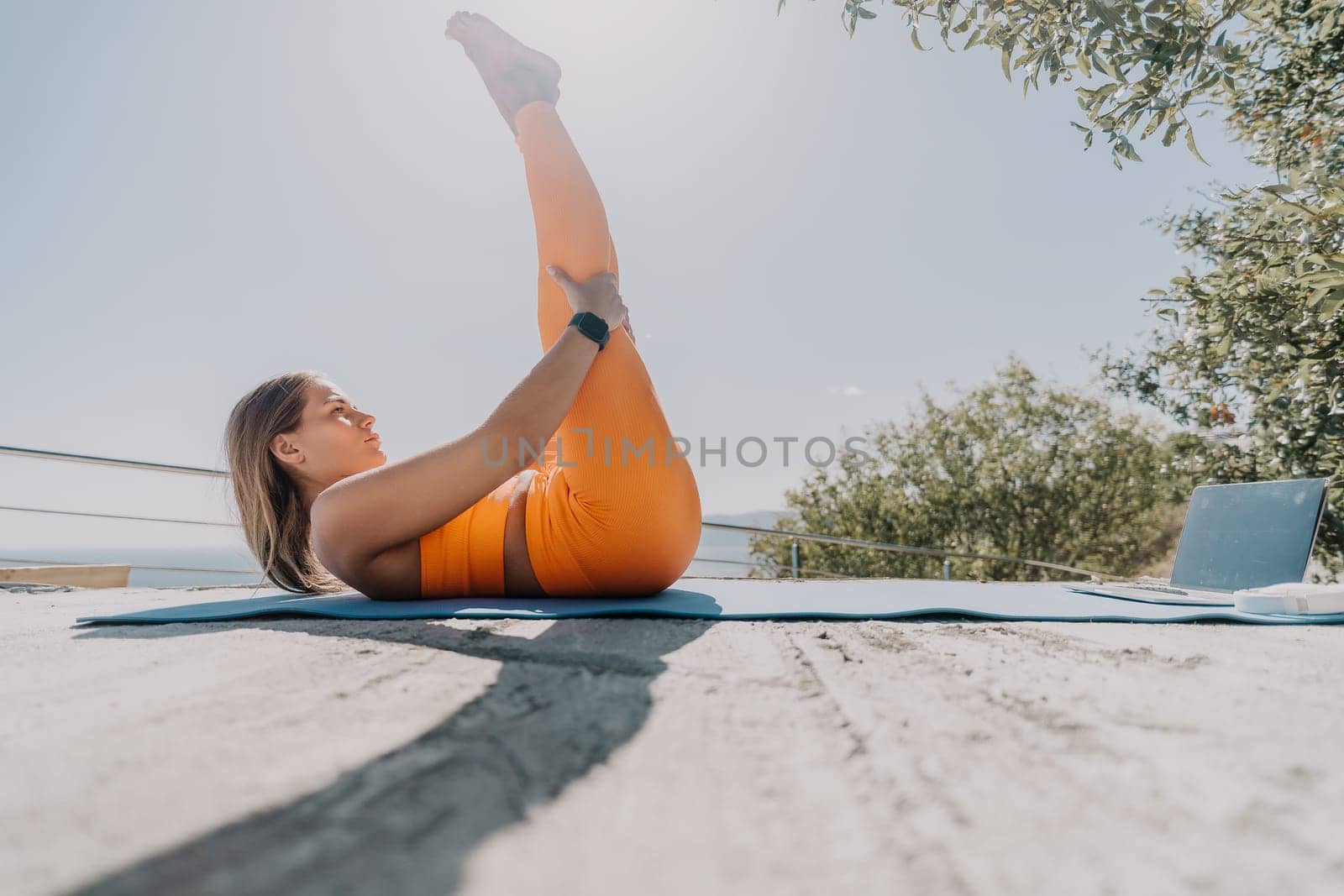 A woman in orange activewear stretches her leg on a yoga mat outdoors with a laptop in the background and a clear sky.