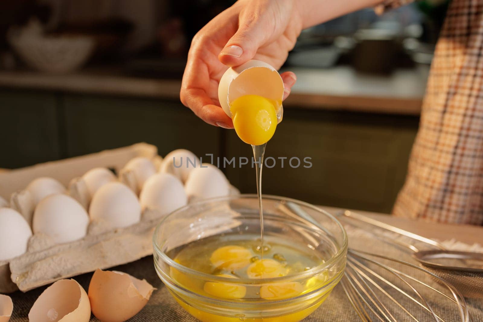 A person cracking an egg into a glass bowl, with several eggs in an egg carton in the background. The scene is set in a kitchen, showcasing the process of preparing food.