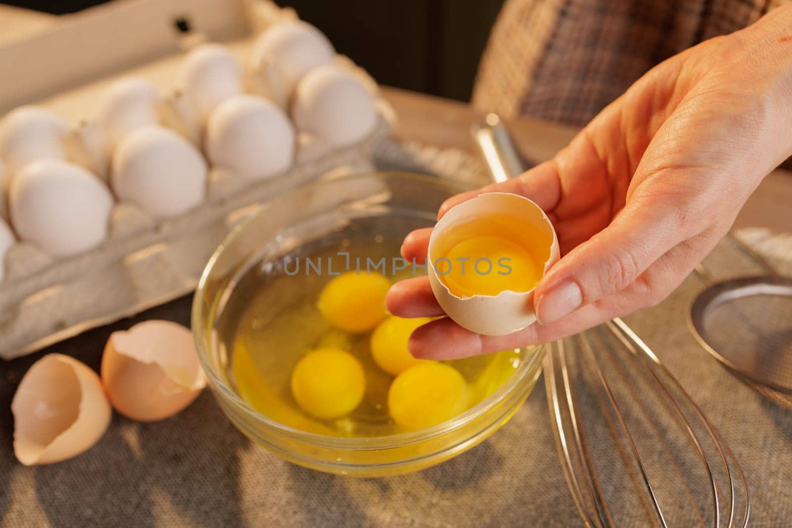 A woman's hand holding a broken chicken egg close-up over a bowl of raw eggs. There are several yolks in the bowl and more eggs in a cardboard box in the background.